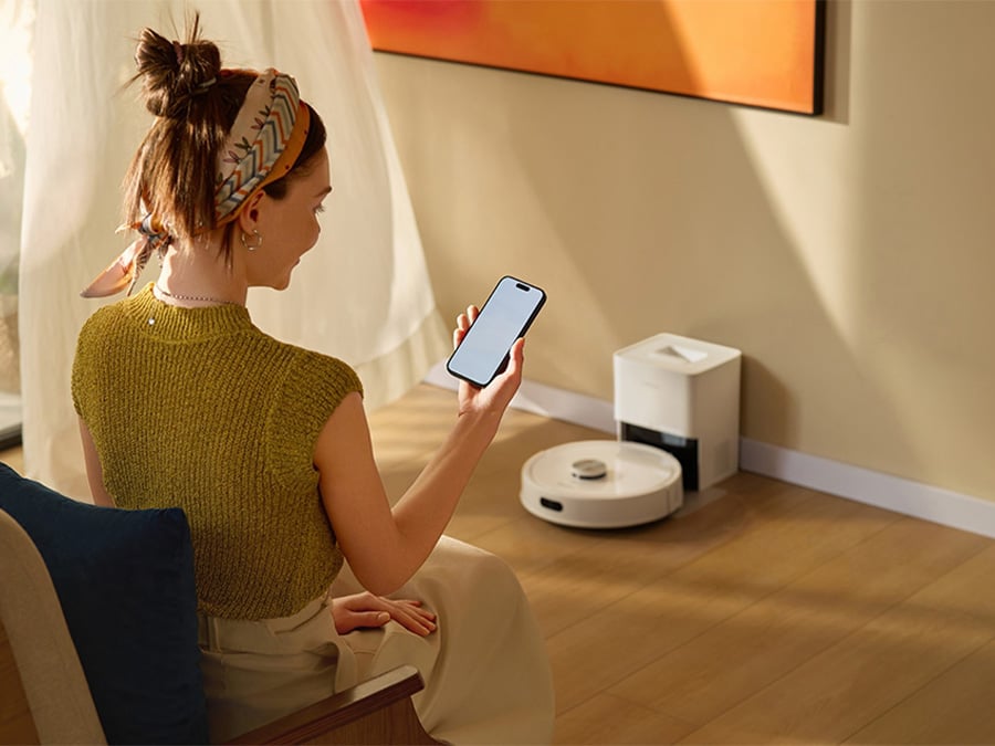 Kobieta siedząca w jasnym salonie steruje robotem sprzątającym Roborock Q7 TF White za pomocą aplikacji mobilnej