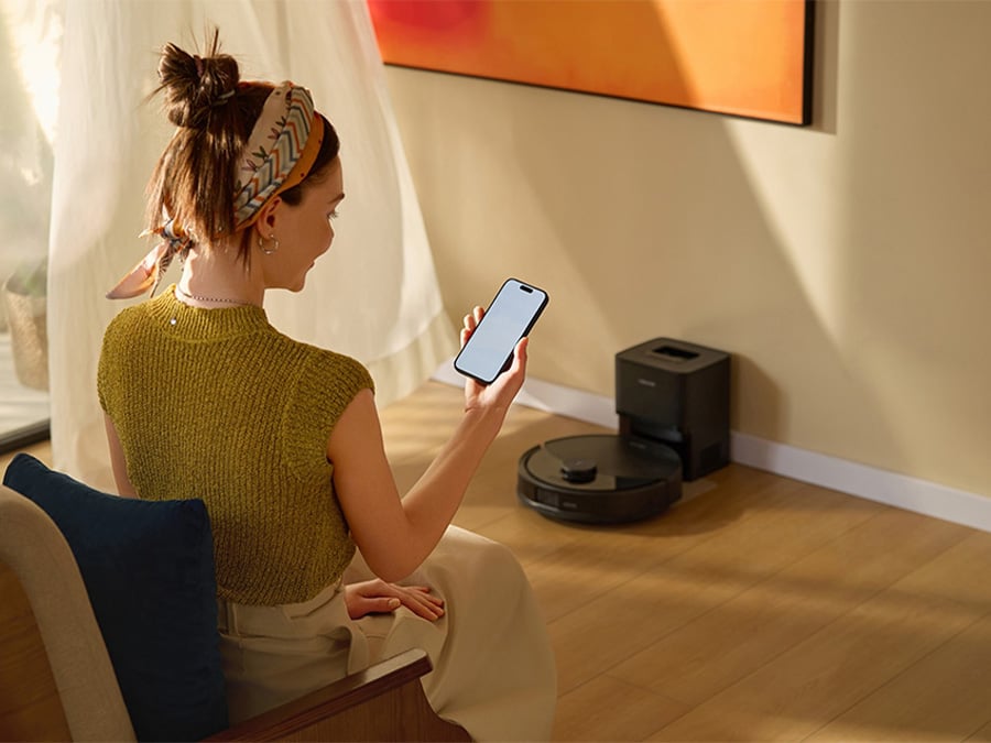 Kobieta siedząca w salonie i obsługująca robota sprzątającego Roborock Q10 za pomocą aplikacji w smartfonie; urządzenie znajduje się przy ścianie w stacji dokującej