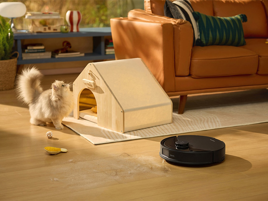 Robot sprzątający Roborock Q10 podczas pracy w salonie, obok bawiący się kot przy legowisku; na podłodze widoczne sierść i zabawki