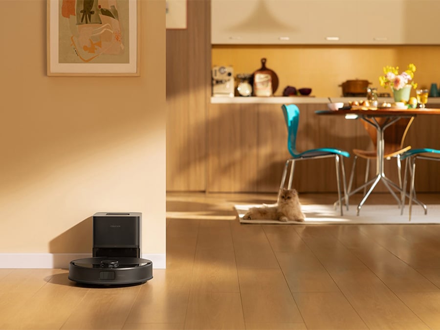 Robot sprzątający Roborock Q10 odkurzający podłogę w pobliżu kota leżącego na dywanie; w tle jadalnia z przygotowanym stołem