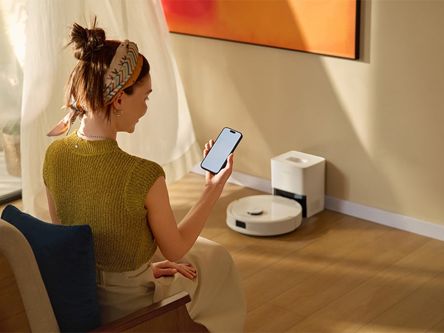Kobieta siedząca w salonie i obsługująca robota sprzątającego Roborock Q10 za pomocą aplikacji w smartfonie; urządzenie znajduje się przy ścianie w stacji dokującej