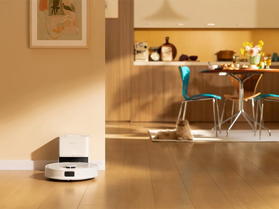 Robot sprzątający Roborock Q10 odkurzający podłogę w pobliżu kota leżącego na dywanie; w tle jadalnia z przygotowanym stołem