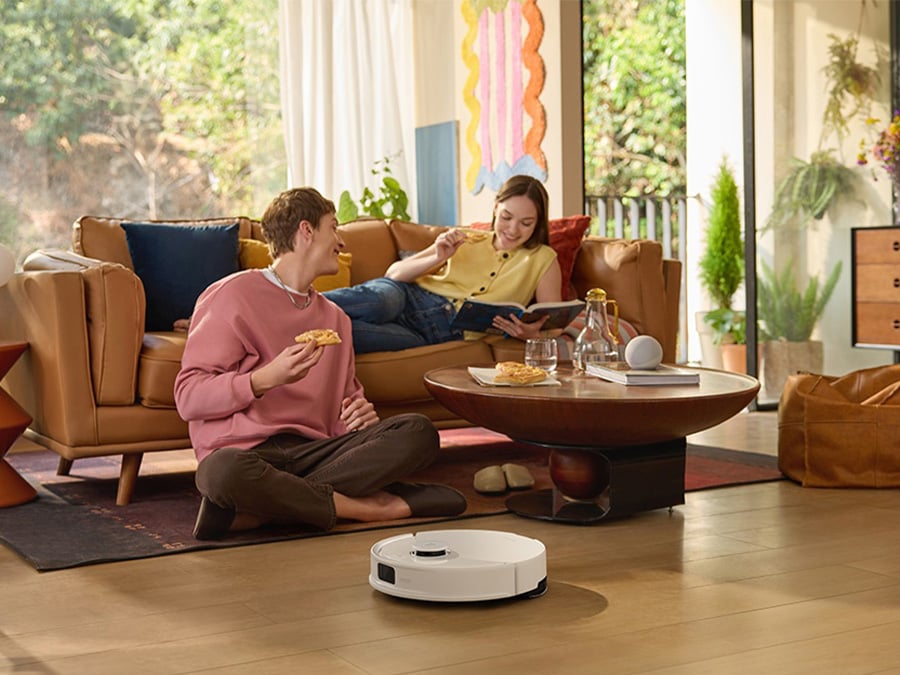 Dwoje młodych ludzi relaksuje się w salonie, podczas gdy biały robot sprzątający Roborock Q10 PF White odkurza podłogę