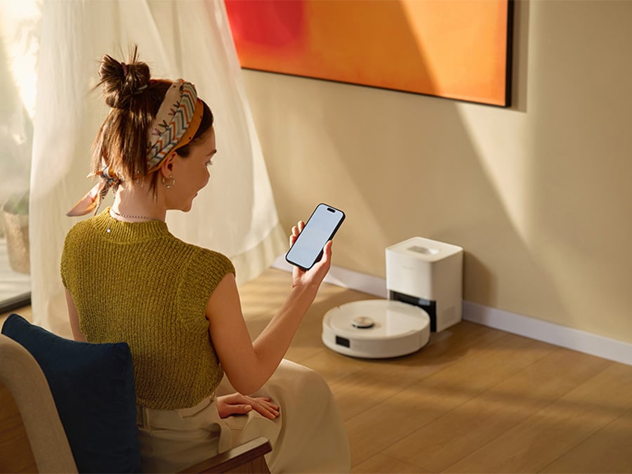 Kobieta siedząca w salonie i obsługująca robota sprzątającego Roborock Q10 VF za pomocą aplikacji na smartfonie