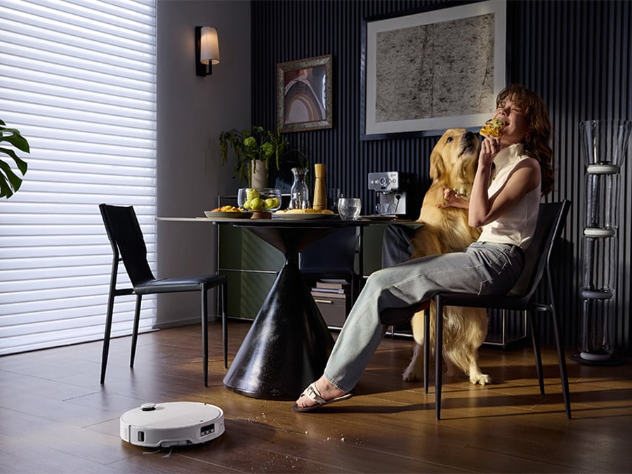 Kobieta jedząca pizzę przy stole w jadalni w towarzystwie psa, a robot sprzątający Roborock Qrevo Edge T White zbiera rozsypane okruchy z podłogi