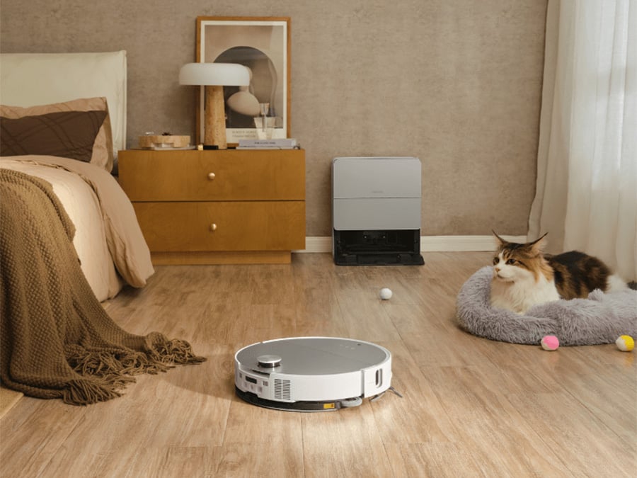Robot sprzątający Roborock Saros 10 White poruszający się po sypialni z leżącym kotem i zabawkami, w tle stacja ładująca