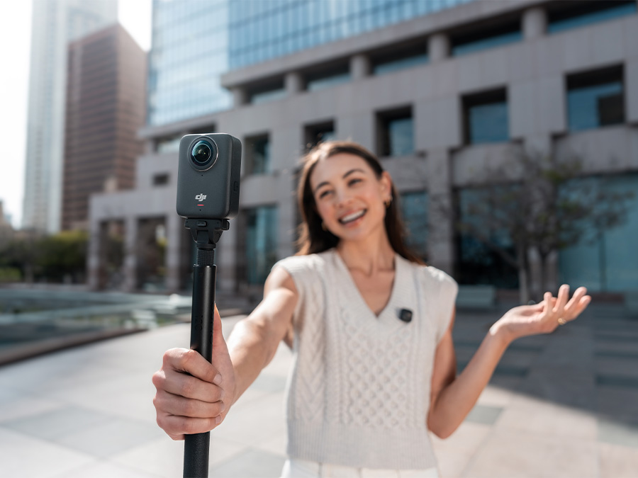 Uśmiechnięta kobieta nagrywająca materiał kamerą DJI Osmo 360 trzymaną na statywie w przestrzeni miejskiej