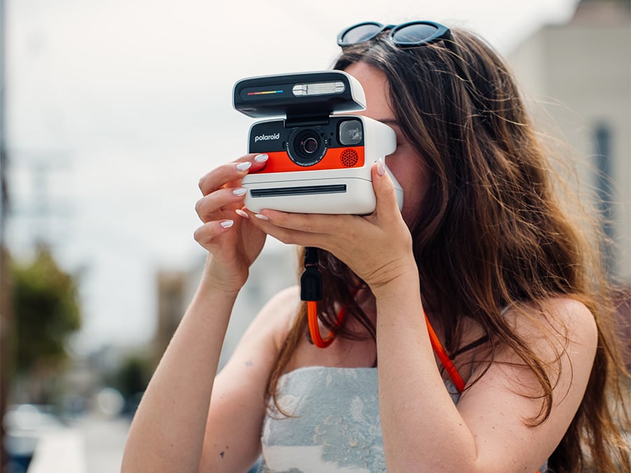 Kobieta robiąca zdjęcie aparatem natychmiastowym Polaroid Flip w białej wersji kolorystycznej z pomarańczowym panelem frontowym i paskiem na szyję