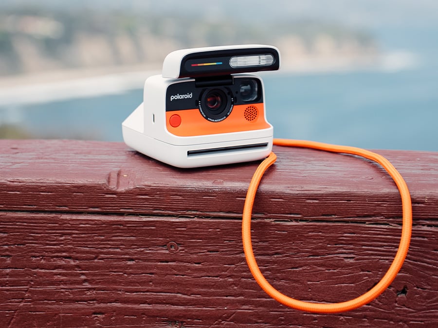 Biały aparat natychmiastowy Polaroid Flip z pomarańczowym frontem i pomarańczowym paskiem na szyję leżący na drewnianej barierce, z widokiem na rozmyte tło nadmorskiego krajobrazu