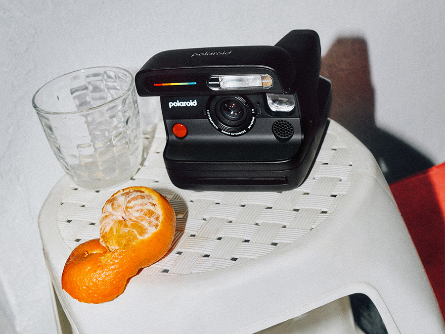 Czarny aparat natychmiastowy Polaroid Flip stojący na białym, plecionym taborecie obok szklanki i obranej mandarynki