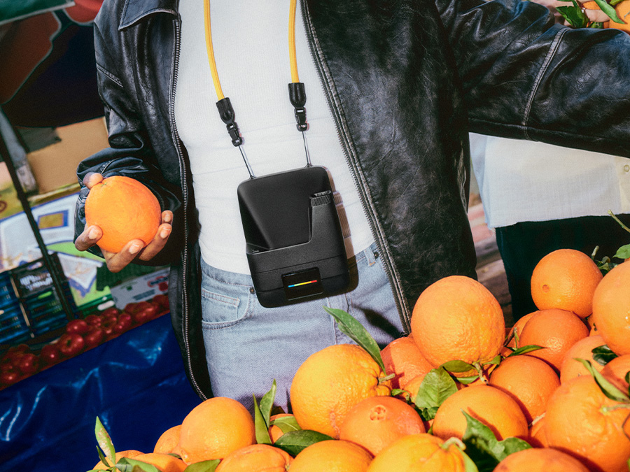 Osoba w czarnej skórzanej kurtce trzymająca pomarańczę na targu, z czarnym aparatem natychmiastowym Polaroid Flip zawieszonym na żółtym pasku na szyi
