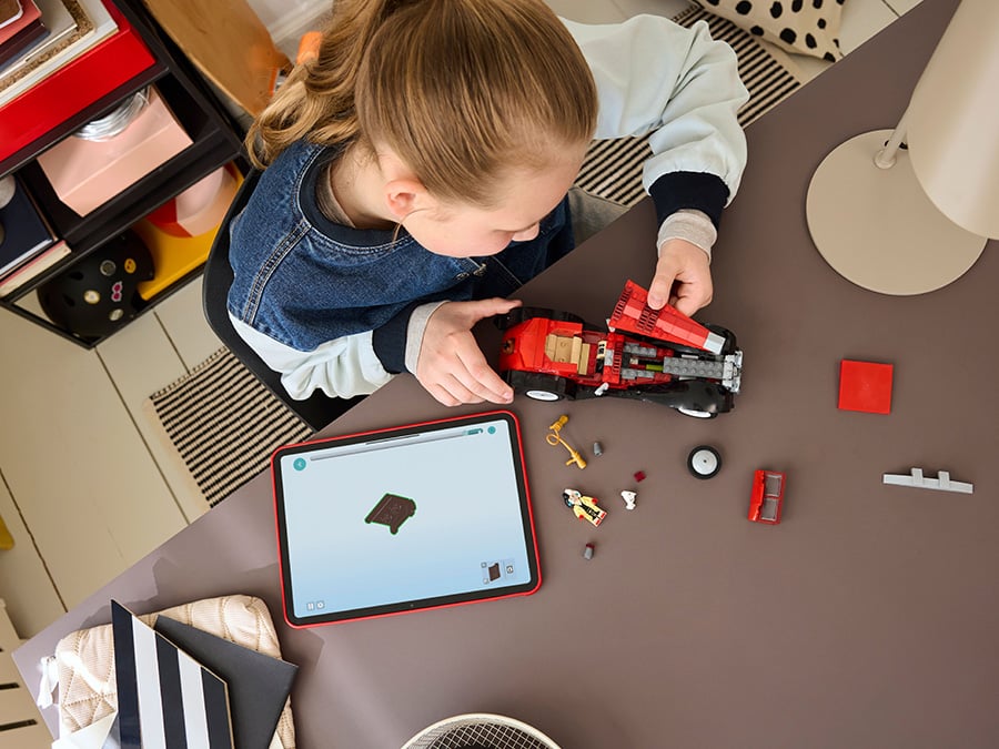 Dziewczynka siedząca przy biurku składa czerwono-czarny samochód LEGO inspirowany Cruellą de Mon, obok tablet z instrukcją budowania i elementy zestawu